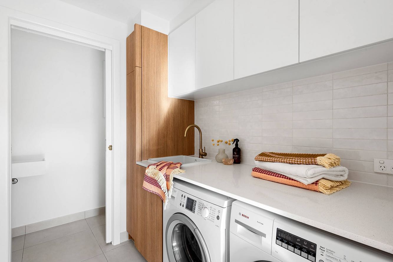 The image depicts a laundry room with white walls and floor, featuring a washing machine and dryer with stacked towels on ...