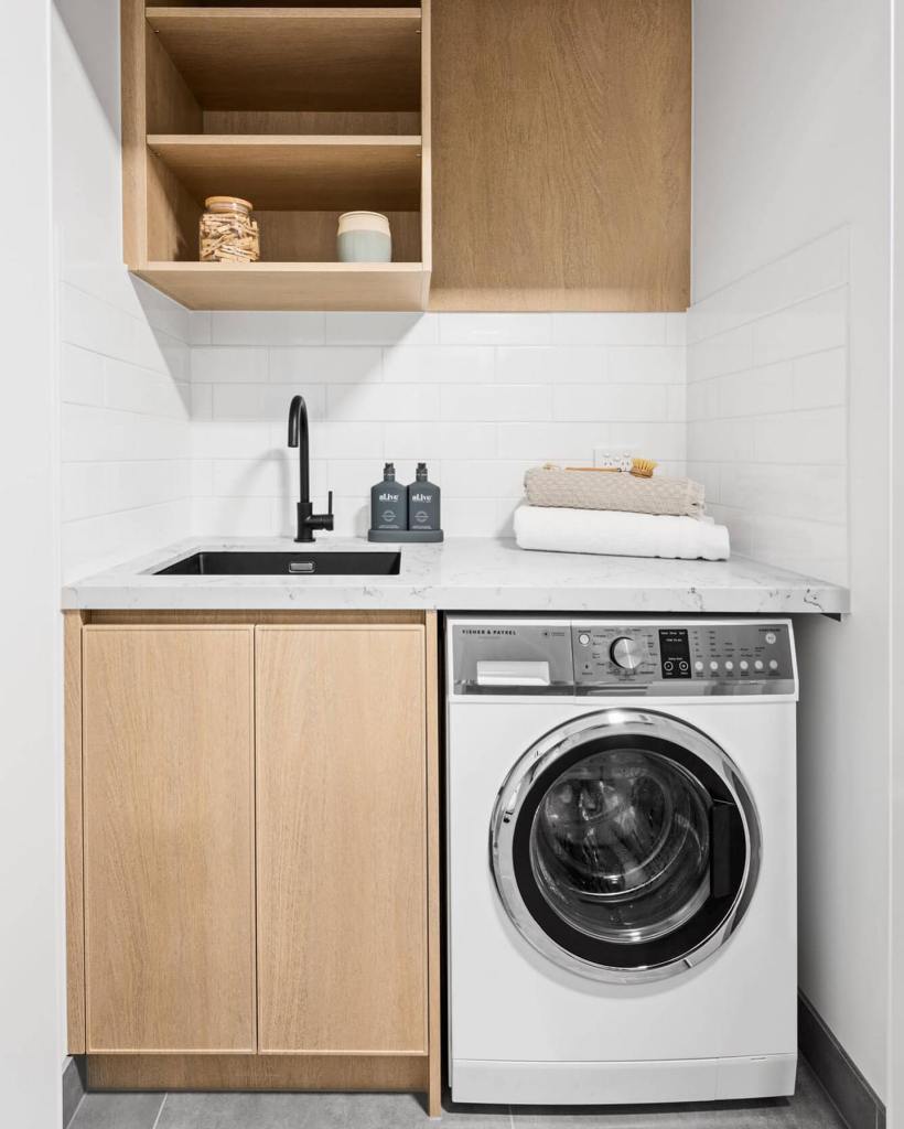 The laundry room features a functional layout, with light wood cabinets and a white marble countertop. A black sink is cen...